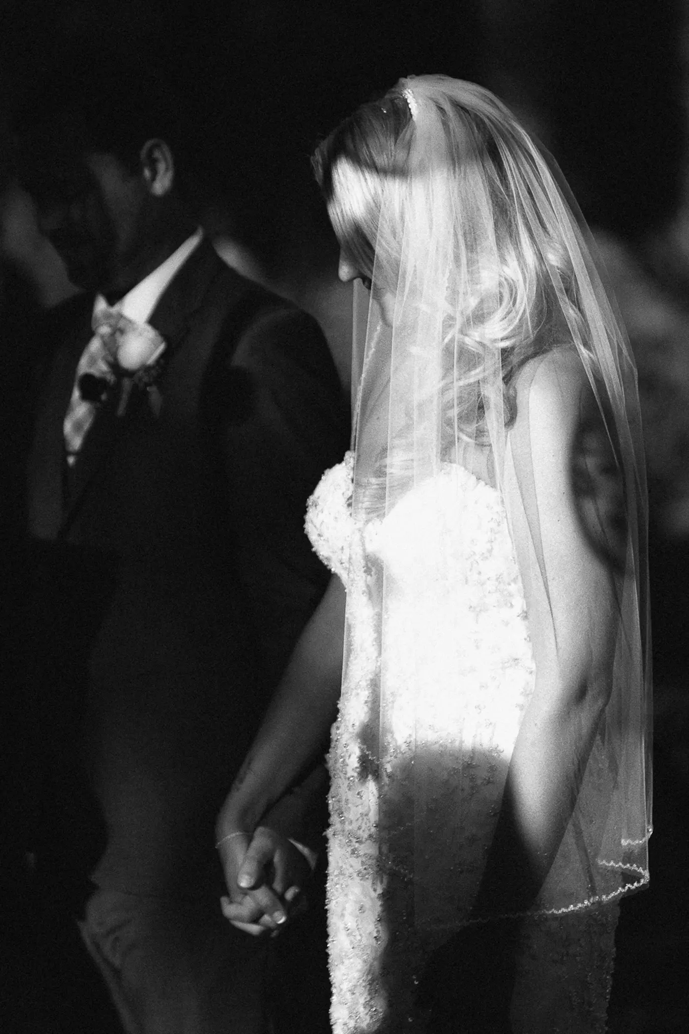 A veiled bride holding hands with her groom in soft black and white light