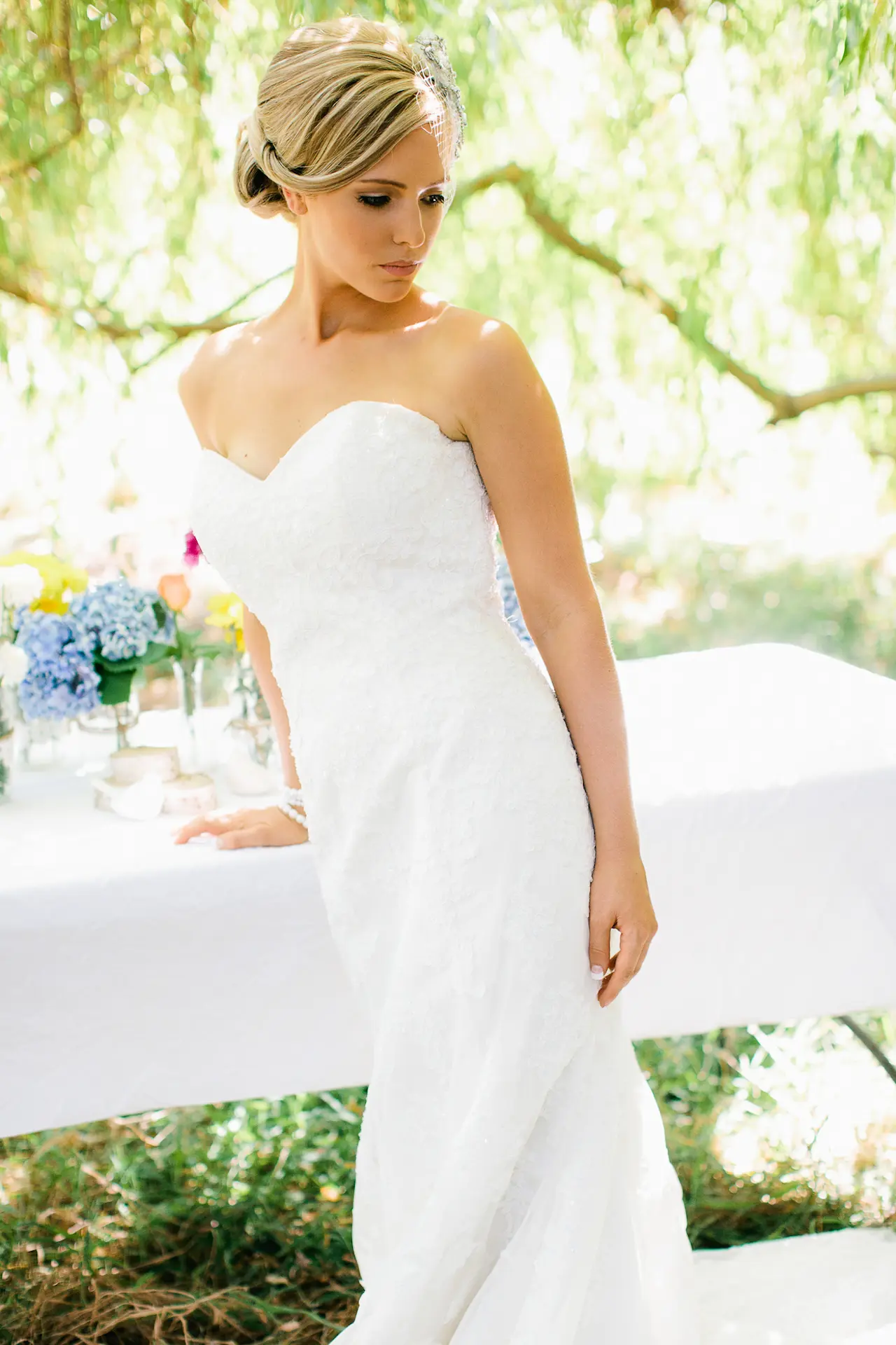 A bride in a strapless gown beneath willow trees beside a flower-laden table